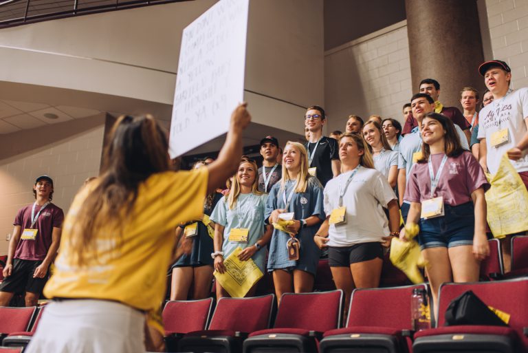 Students at Fish Camp stand in an auditorium while learning Aggie traditions.
