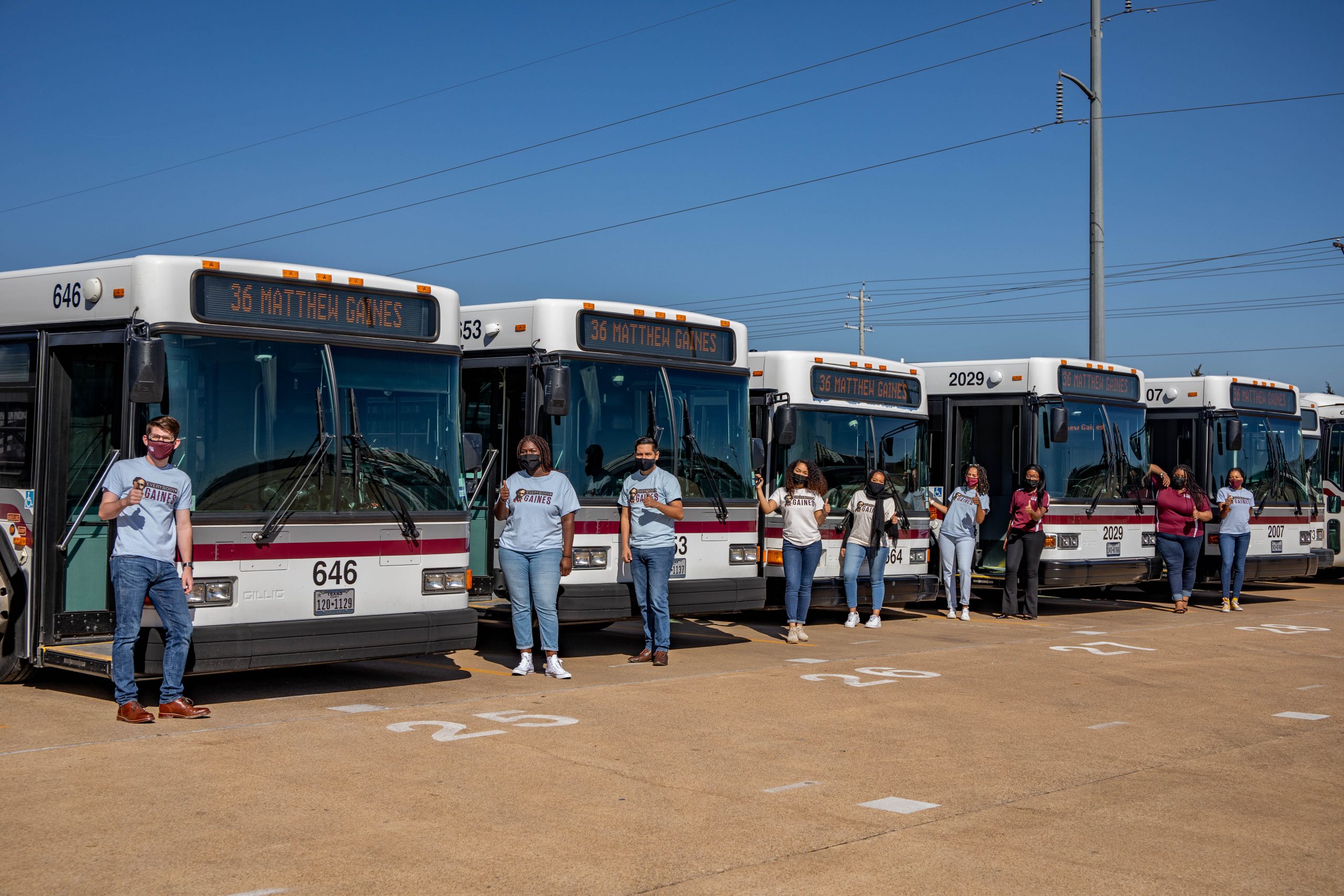 Image of the Matthew Gaines Society in front of a row of Aggie Spirit Buses displaying the newly named Matthew Gaines route on the bus marquees.