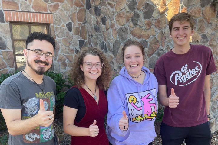 Four Aggie students stand in front of a rock wall giving thumbs up.