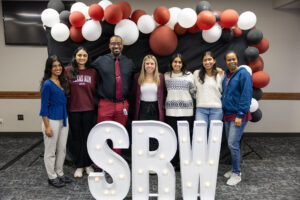 the student research week planning team posing in front of a photo backdrop with balloons and big letters that spell SRW
