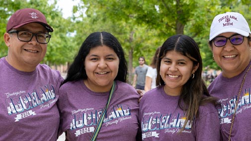 Two students with their parents smiling for a photo in Aggie Park.