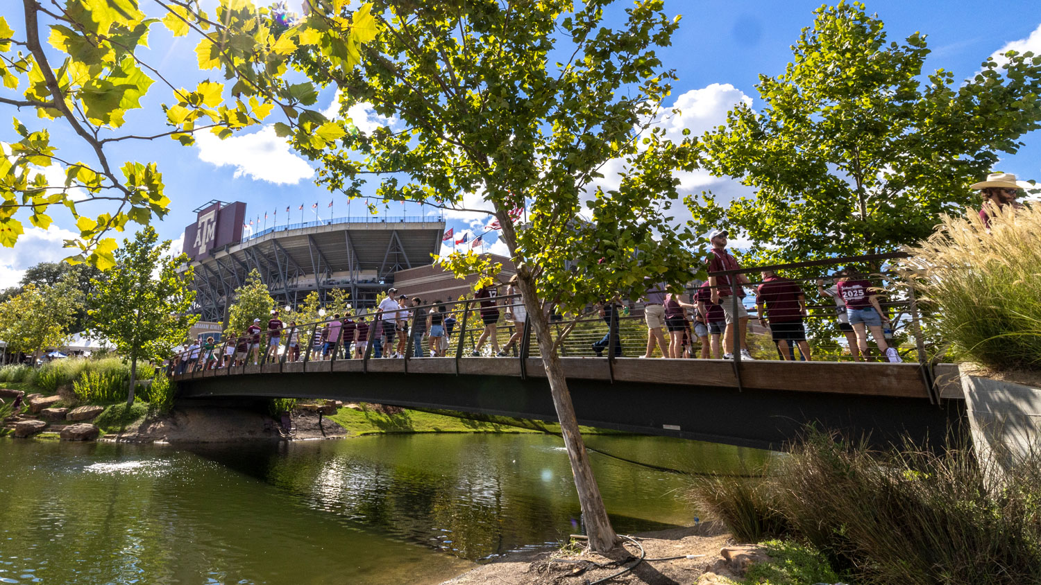 Aggie Park bridge over the pond filled with people with Kyle Field in the background.