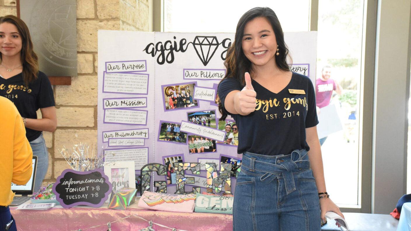 Student giving a Gig 'Em in front of an Aggie Gems display in the MSC.